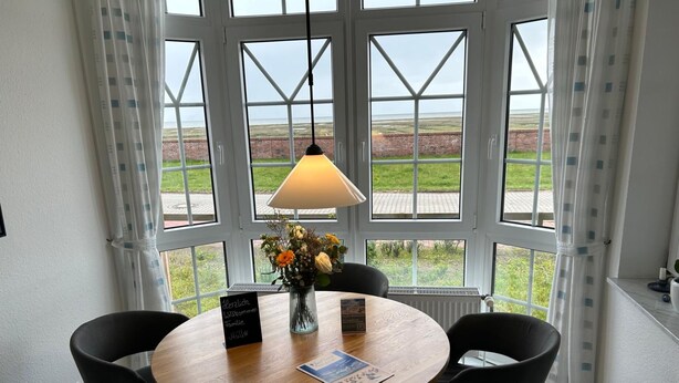 Ein Zimmer mit einem runden Holztisch, vier schwarzen Stühlen, einer Blumenvase, einer Tischlampe und Blick auf ein grünes Feld durch große Fenster.