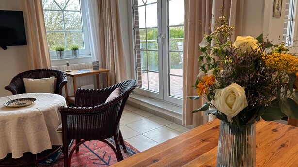 Ett rum med ett träbord, flätade stolar, en vit duk, en vas med blommor och stora fönster med gardiner.