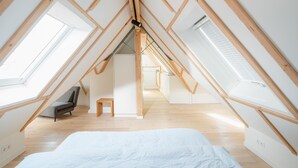 Un dormitorio abuhardinado con altas paredes, tragaluces, una cama, una silla y una mesita.