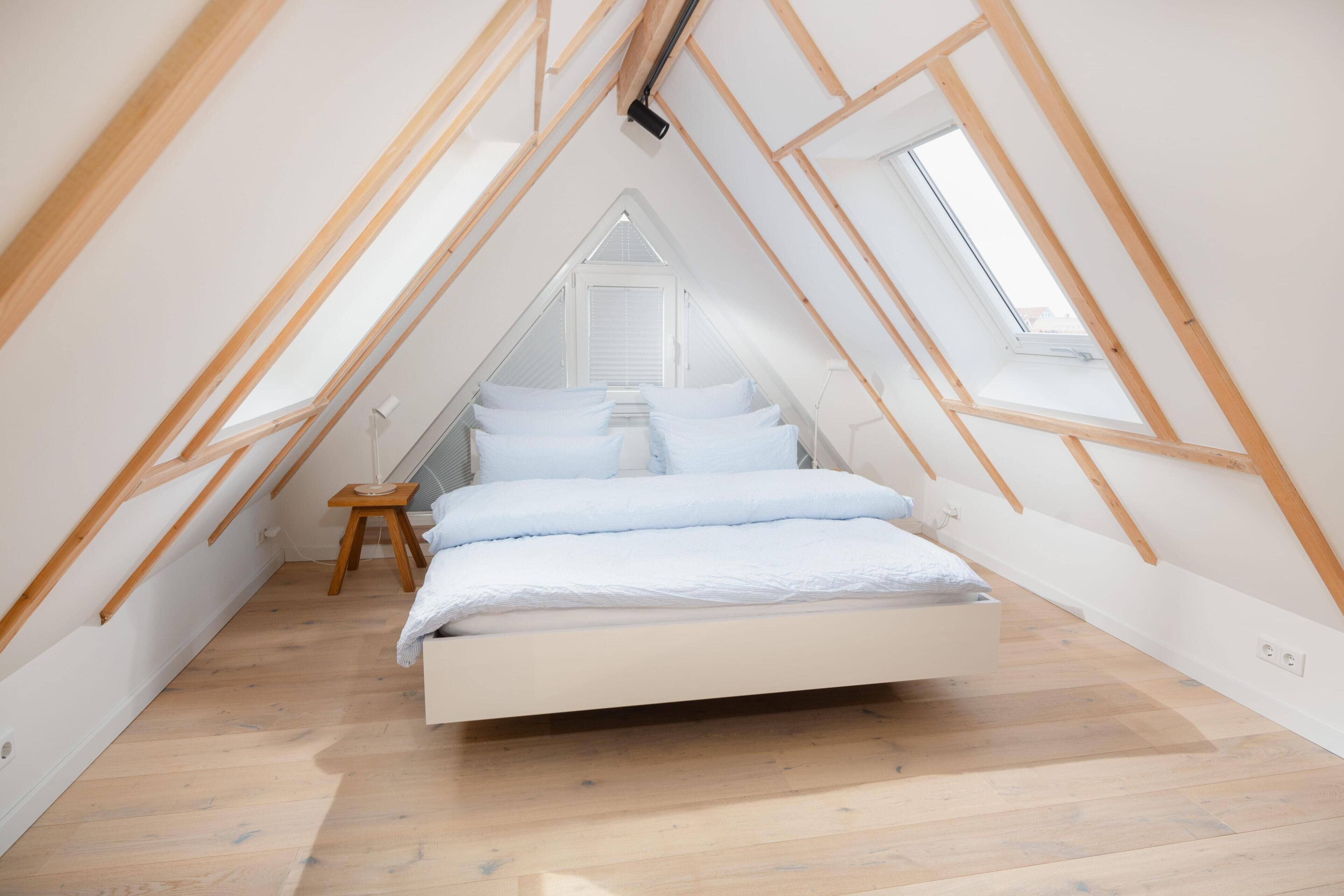 Une chambre avec un plafond en pente, un lit, un petit tabouret et une fenêtre.