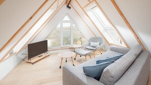 Un dormitorio tipo loft moderno con techo inclinado, un ventanal grande, un televisor de pantalla plana, un sofá gris, una mesita redonda y una silla.
