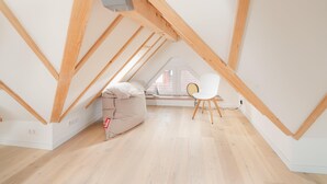 Habitación en ático con techo inclinado, vigas de madera, una silla blanca, un sillón de peluche y una ventana con vistas.