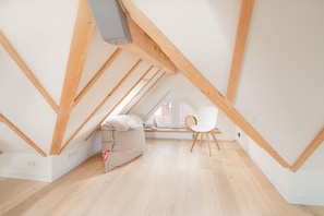 Ein Dachzimmer mit schrägem Dach, Holzbalken, einem weißen Stuhl, einem Sitzsack und einem Fenster mit Ausblick.