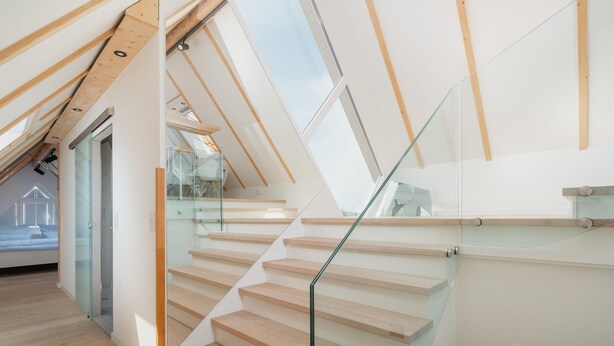 Un interior moderno con escalera de madera, barandas de vidrio y techo inclinado.