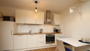A modern kitchen with white cabinets, a wooden countertop, a built-in oven, a coffee maker, and a dining area with a white table and blue chairs. - Vacation apartment Bootjefahrt at Uns to Hus (Juist)