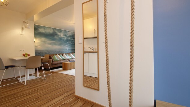 A modern room with a wooden floor, a dining area with chairs, a sofa with cushions, a large mirror, and a rope wall decoration. - Vacation apartment Bootjefahrt at Uns to Hus (Juist)