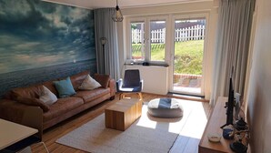 A living room with a brown leather sofa, a wooden coffee table, a blue chair, a small ottoman, a TV, and a large window with a view of a white fence and greenery. - Vacation apartment Bootjefahrt at Uns to Hus (Juist)