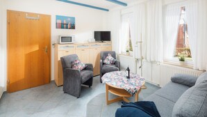 Olohuone, jossa on sohva, kaksi nojatuolia, kukkapuvukuvioitu pöytäliina, televisio, mikroaaltouuni ja puinen ovi.