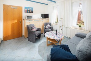 A living room with a sofa, two armchairs, a table with a floral tablecloth, a television, a microwave, and a wooden door. - Frauke apartment in the Villa Altmanns (Juist)