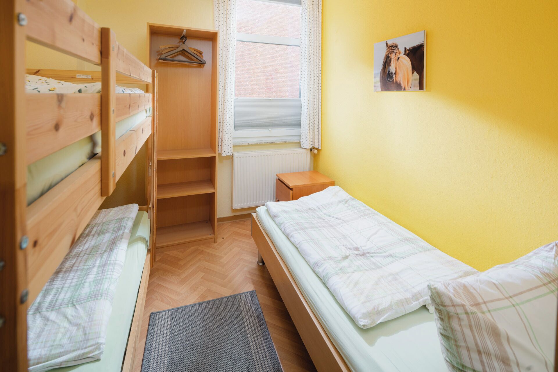 Et værelse med køjesenge, et lille natbord, en radiator og et vindue med gardiner.