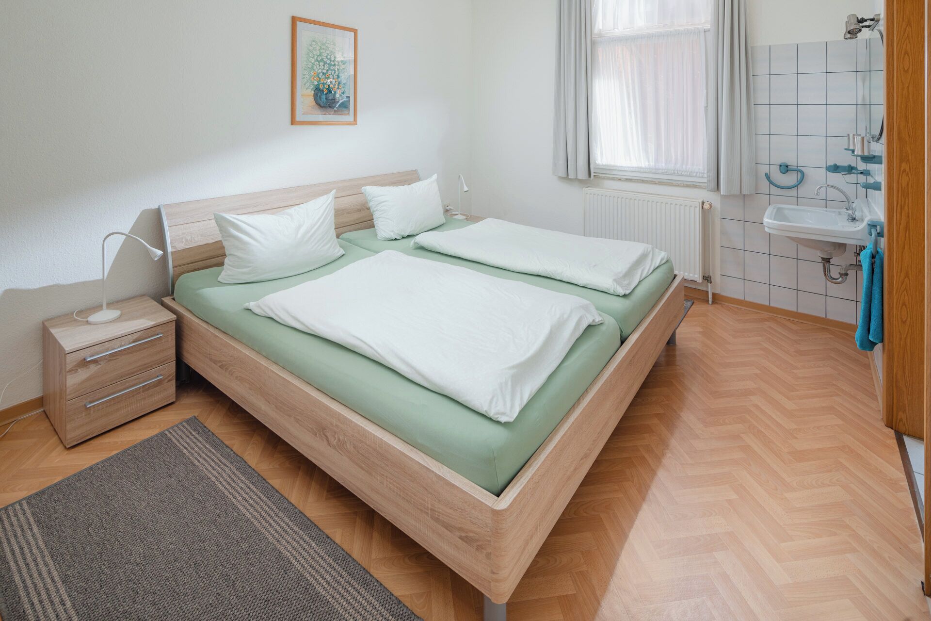 Una camera da letto moderna e pulita con un letto di legno, un comodino, un termosifone, una finestra con tende e un bagno con lavandino e portasci.