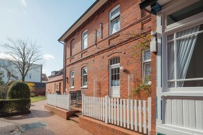 Une maison à deux étages, construite en briques, avec une clôture en bois peinte en blanc, un petit porche à l’avant et une porte de garage bien visible.