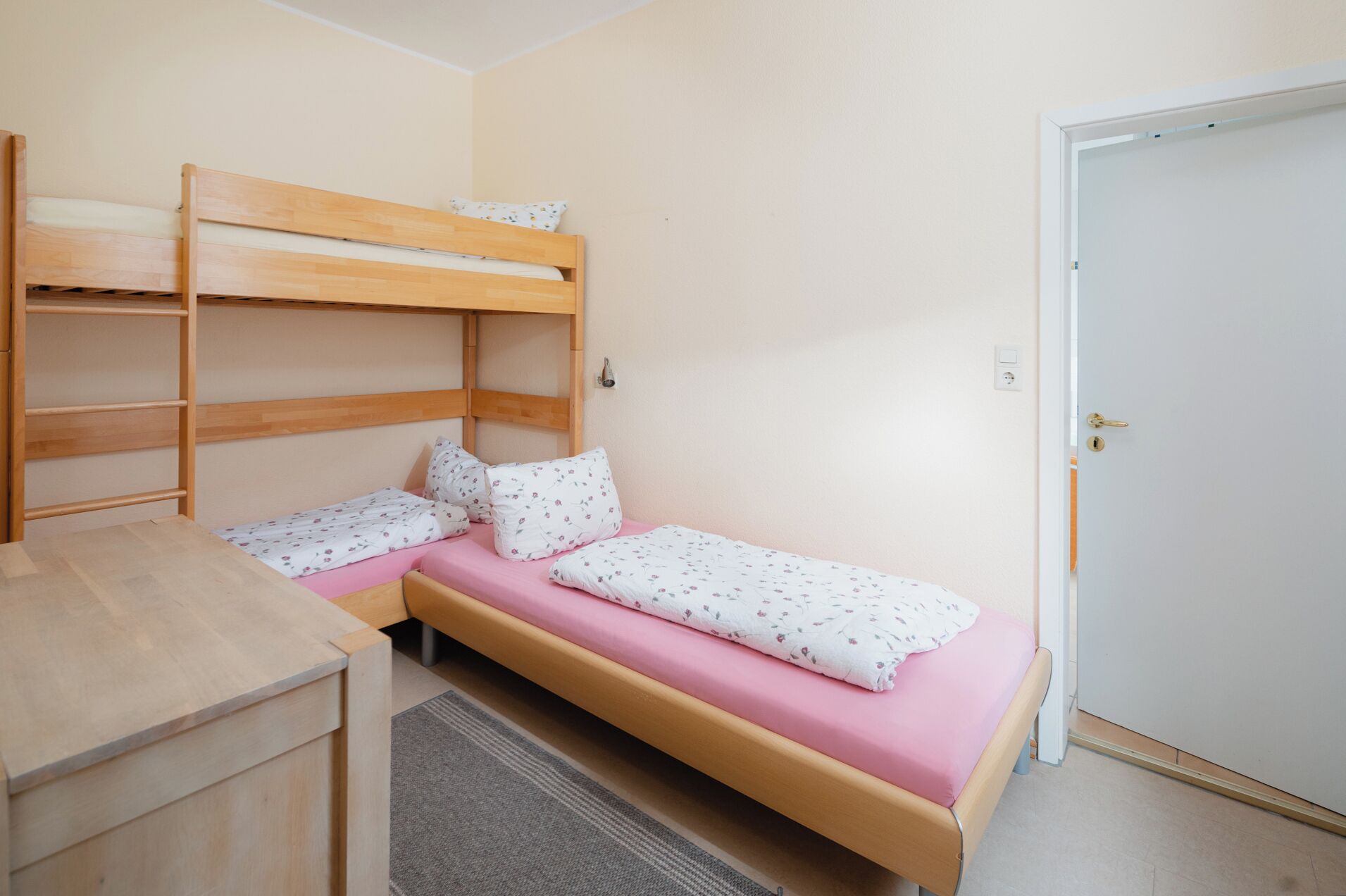 A single bed with a pink mattress and floral bedding, a wooden bunk bed to the left, a small wooden desk, and a door to the right.
