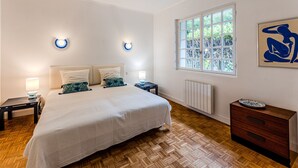 Une chambre à coucher comprenant un lit, des tables de chevet, un radiateur, une fenêtre et une œuvre d’art.