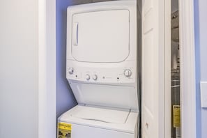 Eine weiße Waschmaschine steht in einer Ecke des Raumes, in der Nähe einer Tür und einem sichtbaren Wasserrohr.