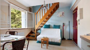 A room with a wooden staircase, a bed with teal bedding, a dining table set with plates and glasses, and a window with a view of trees. - Duplex in Carolles, Bay of Mont Saint Michel, 50 m from the Beach (Carolles)
