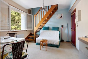 Une piĂšce avec un escalier en bois, un lit avec une literie de couleur turquoise, une table Ă manger dressĂ©e avec des assiettes et des verres, et une fenĂȘtre donnant sur des arbres.