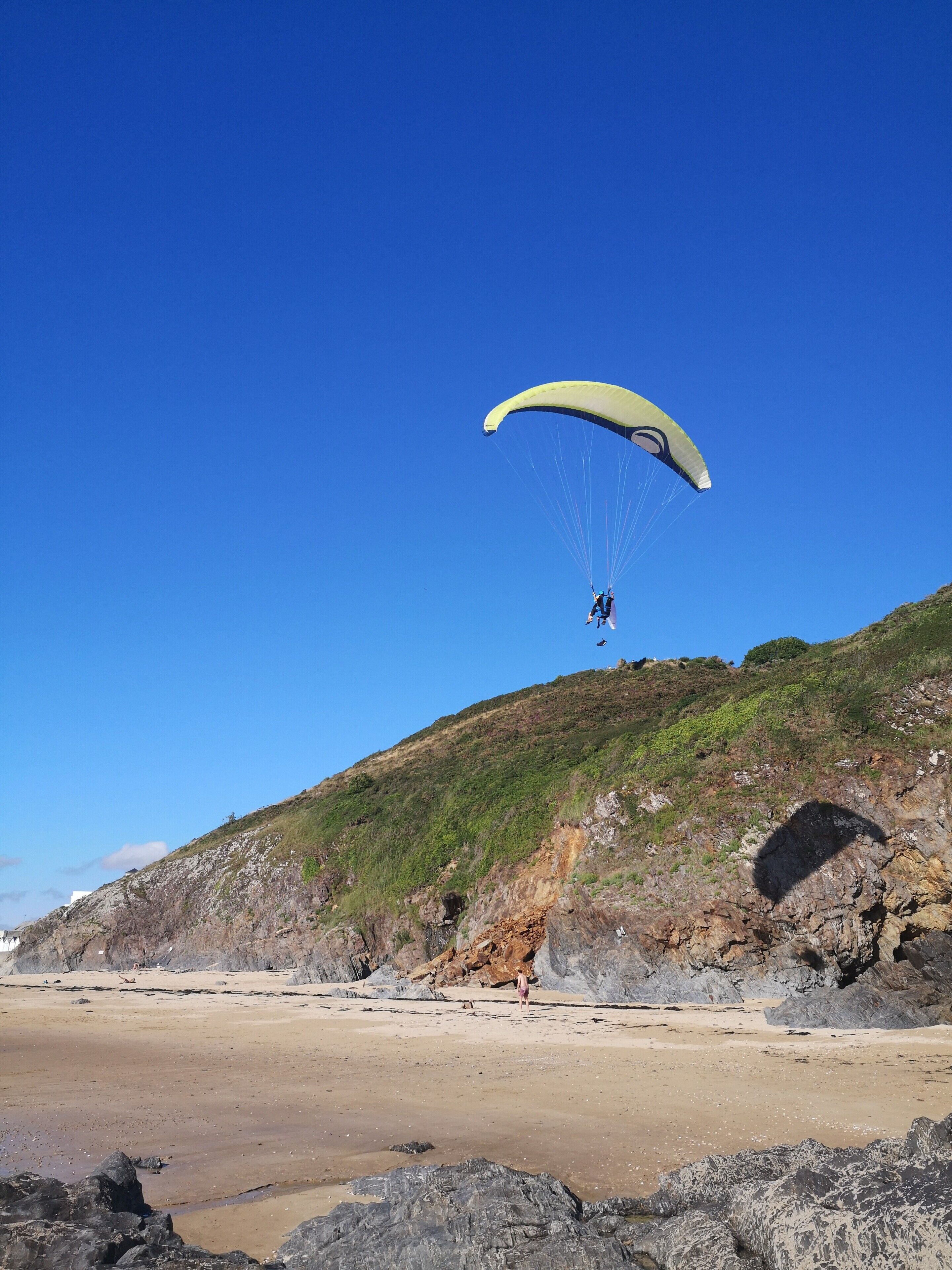 Ein Strand mit einer Person, die Gleitschirm fliegt, felsigem Gelände und blauem Himmel.