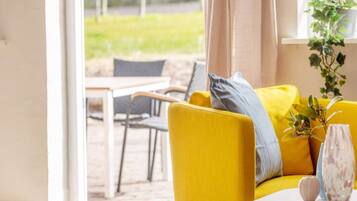 A bright yellow sofa with a grey pillow, a white vase with flowers, and a potted plant on a windowsill.