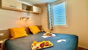 Ein Schlafzimmer mit einem Bett, einem Frühstücksbrettchen, einem Fenster mit Jalousien und einer hängenden Pflanze.