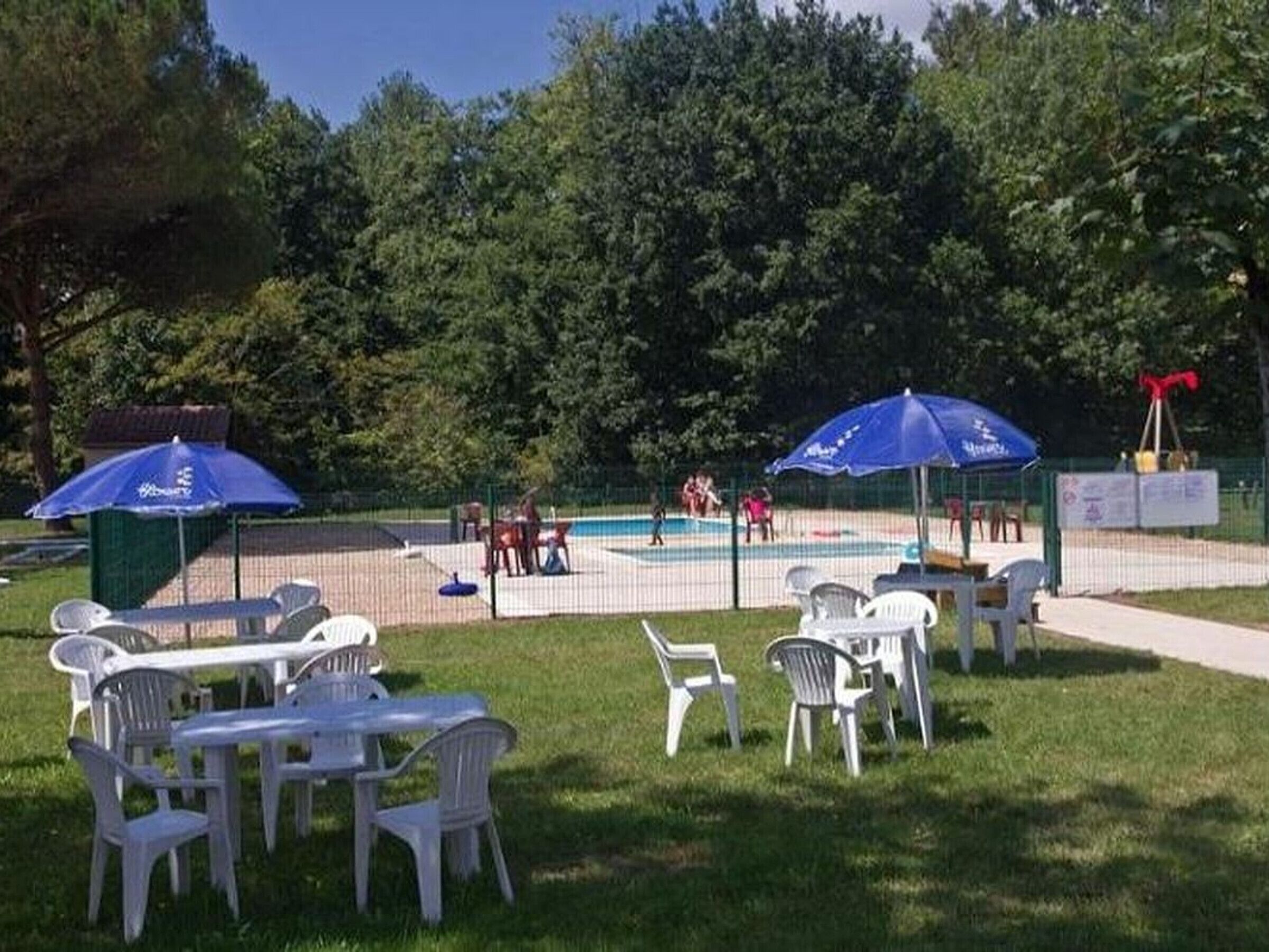 An outdoor pool area with a volleyball net, surrounded by white plastic chairs and tables, and shaded by blue umbrellas.