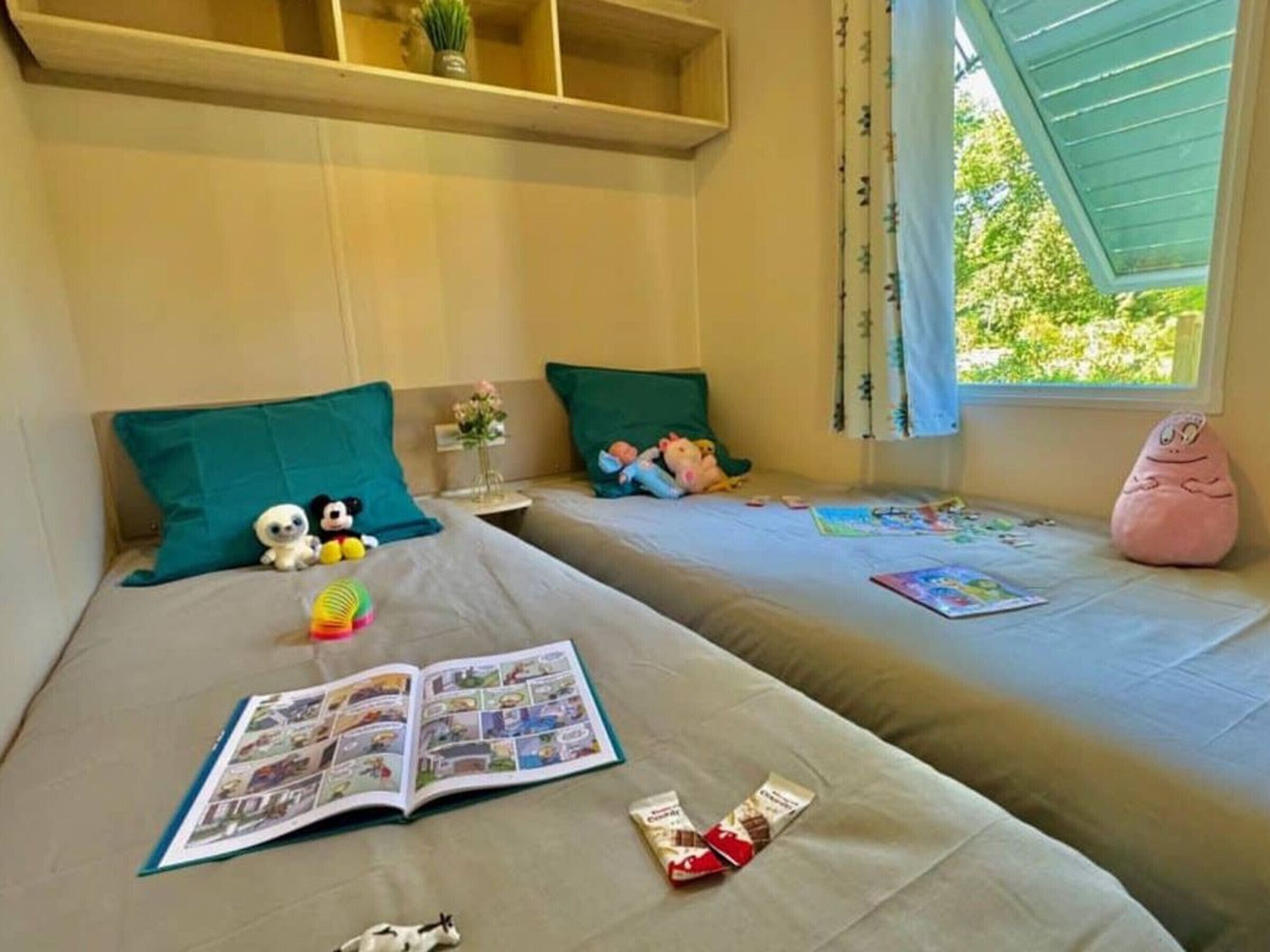 A room with two beds, each with a pillow and stuffed toys. There is a window with curtains, a shelf above the beds, and a book on the floor.