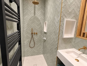 Un baño moderno con un salpicadero de baldosas tipo espiga, una ducha con cabezal de ducha de mano, un lavamanos blanco con grifo dorado y un perchero para toallas.