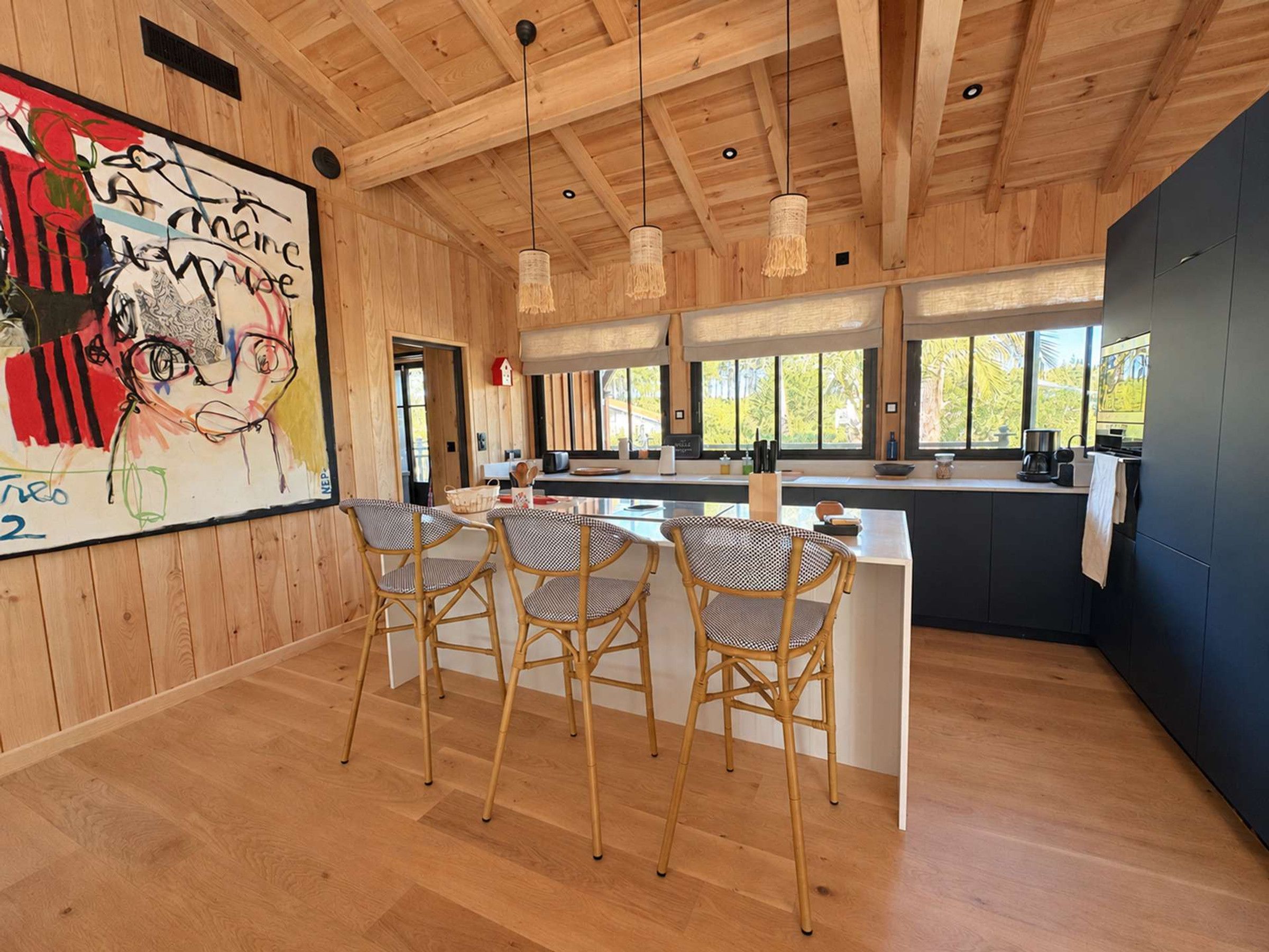 A modern kitchen with a bar area, wooden ceiling, and large windows.