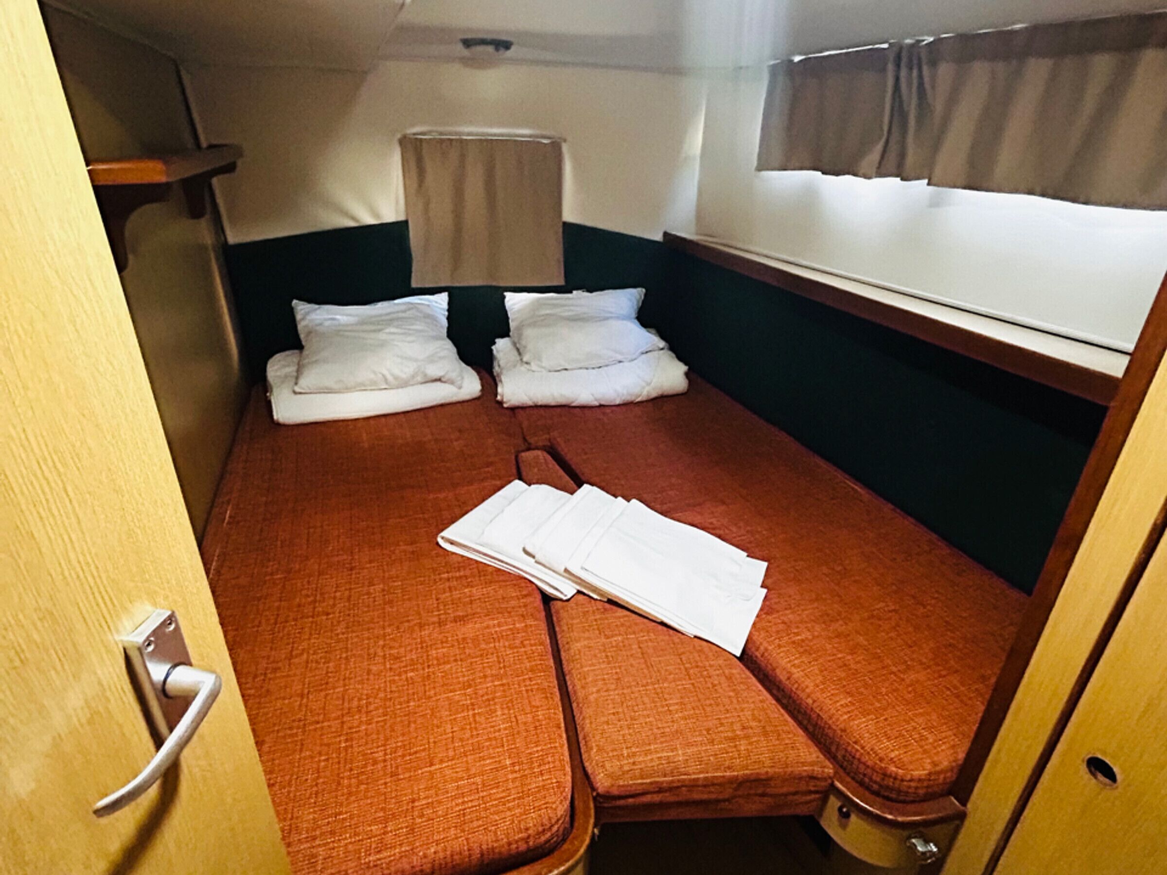 A compact, single-bed room with a wooden door, a small window with curtains, and a simple bed with pillows and a folded blanket.