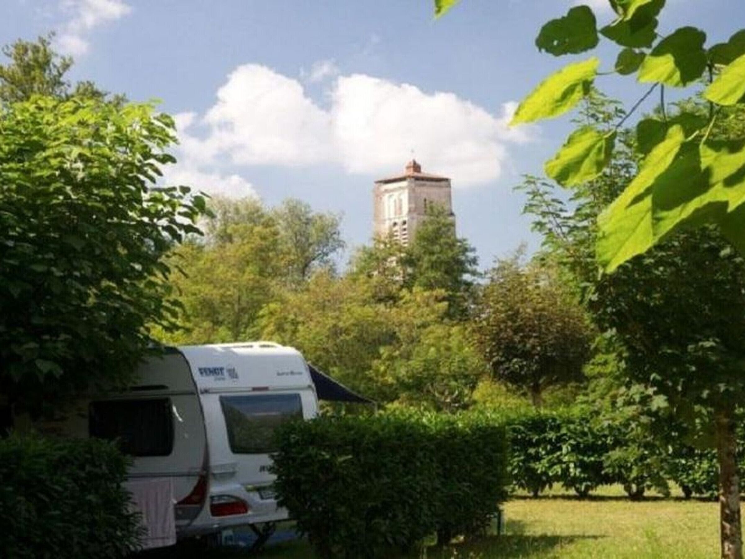 Ein weißer Wohnmobilwagen steht auf einer Wiese mit Bäumen und einem Turm im Hintergrund.