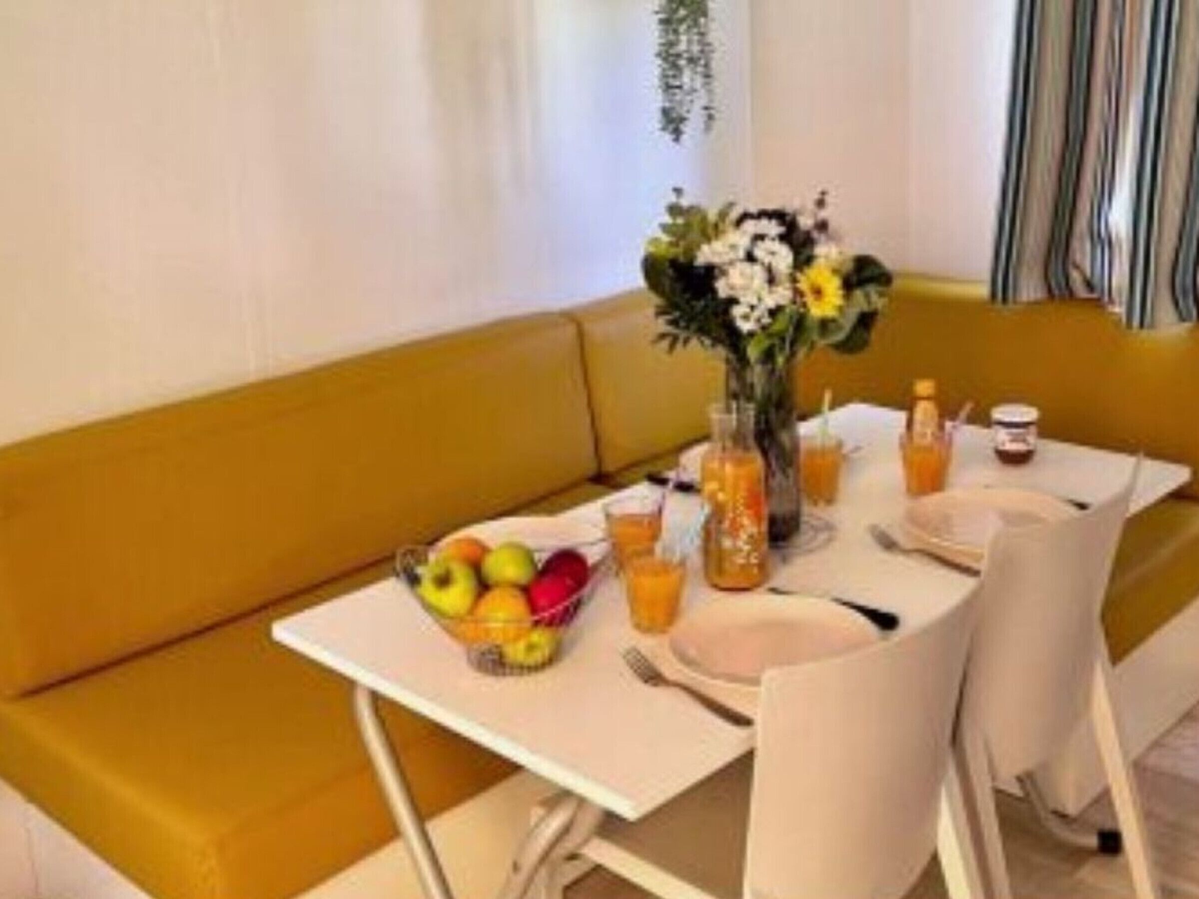 A dining area with a table set for two, a bowl of fruit, a vase with flowers, and a bottle of orange juice.