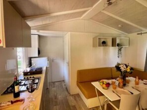 A compact kitchen with a stove, sink, and microwave. There is a dining area with a table set for two, a bench, and a vase with flowers. - Mobile home with terrace - 5 guests (Saint-Astier)
