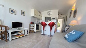 Una sala de estar con un sofá, un televisor y una estantería. Hay una zona de cocina con un reloj y un comedor con sillas rojas.