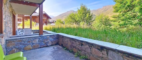 A covered outdoor seating area with stone walls, a green plastic chair, and a view of mountains and greenery.