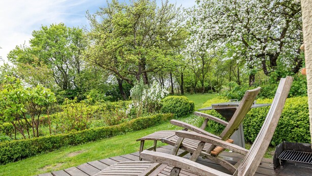 A wooden deck with outdoor seating, surrounded by lush greenery and flowering trees.