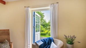 A room with a wicker chair, a small table with a book, a potted plant, and a view of a garden through an open door.