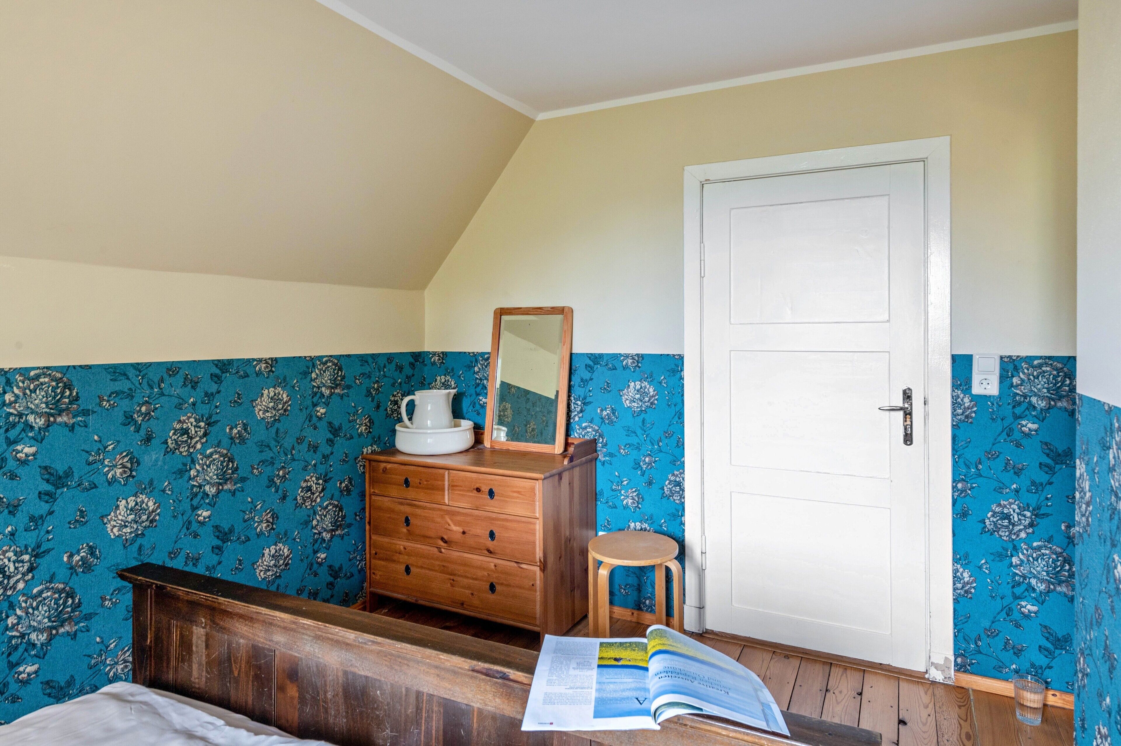 A bedroom with a wooden dresser, a mirror, a stool, and a bed with a book on it.