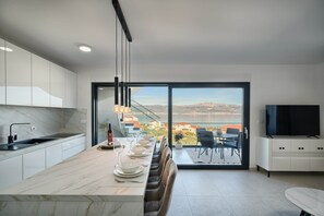 A modern kitchen with a marble countertop, a dining area with a long table, and a view of the sea through large glass doors.