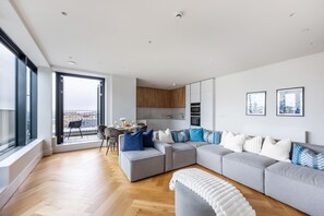 Ein modernes Wohnzimmer mit einer grauen Eckcouch, einem weißen Hocker und einem Essbereich mit Tisch und Stühlen.