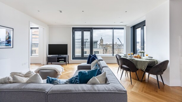 Una sala de estar moderna con sofá, televisor, mesa de comedor y ventanas amplias que ofrecen vista a la ciudad.