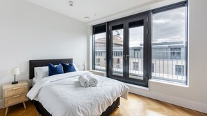 Un dormitorio moderno con una cama grande, una mesita de noche, una lámpara y vistas a edificios a través de amplias ventanas.