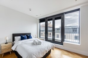 Un dormitorio moderno con una cama grande, una mesita de noche, una lámpara y vistas a edificios a través de amplias ventanas.