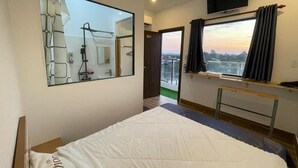 A hotel room with a bed, a large mirror, a TV mounted above the bed, a window with curtains, and a view of the cityscape. - Ngoc An Hotel (Long Hoa)