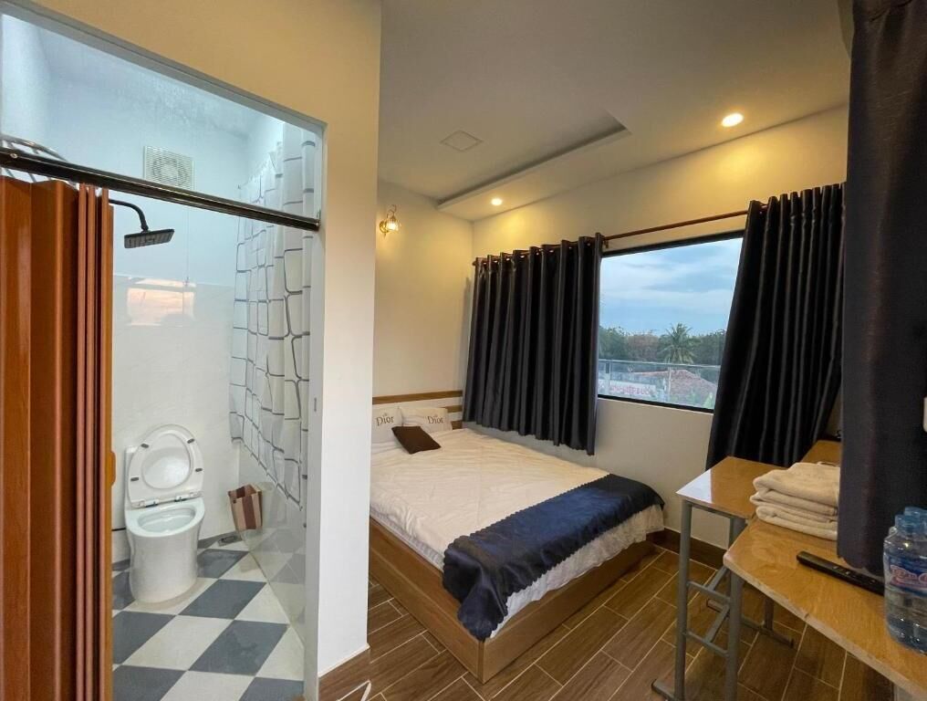 Ett hotellrum med en säng, en dusch, ett fönster med gardiner, ett bord med en flaska och en incheckningsdisk.