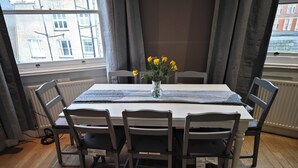 Ein Esstisch mit Stühlen, eine Vase mit gelben Blumen und Blick auf Gebäude durch ein Fenster.