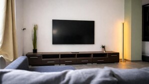 A modern living room with a flat-screen TV mounted on the wall, a dark wooden entertainment unit, a floor lamp, and a grey sofa. - Modern SPA studio in the heart of Gehrden (Gehrden)