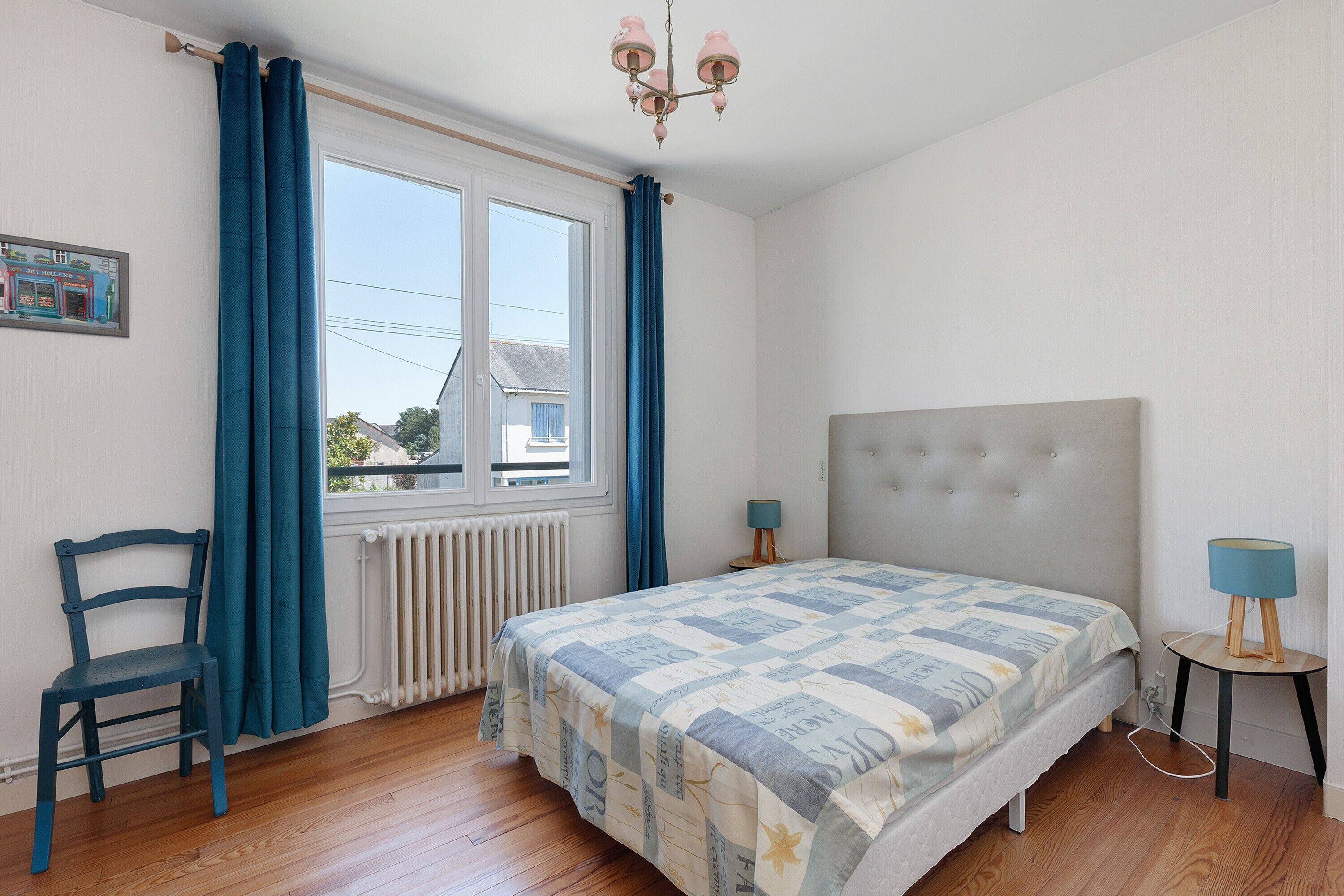 A bedroom with a bed, a chair, a window with curtains, a radiator, and a chandelier.