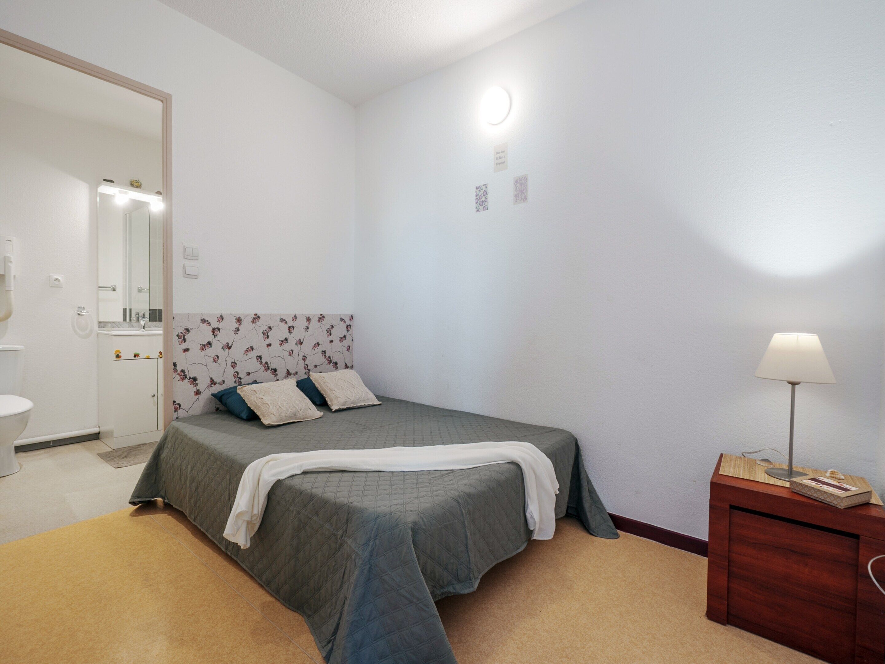 A bedroom with a bed, a bedside table, a lamp, and a bathroom visible in the background.