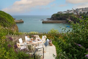 A coastal outdoor seating area with a table, chairs, and a view of the sea and houses in the background. - Little Northcliffe is uniquely situated on the clifftop overlooking Port Isaac (Port Isaac)