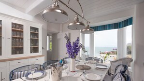 Spiseområde med et langt bord dækket til fire personer, omgivet af fletstole, et hvidt skab med glaslåger og udsigt til havet.